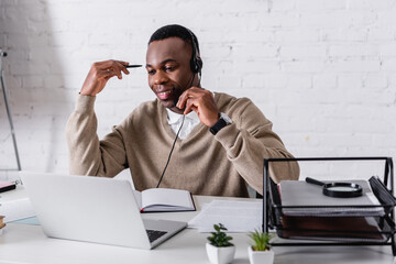 smiling african american interpreter in headset holding pen while working at laptop in translation...