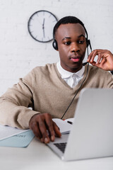 young african american translator in headset working at laptop on blurred foreground