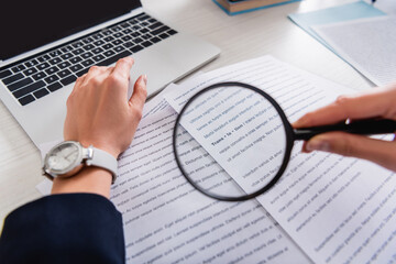 cropped view of translator holding magnifier glass near documents with english text, blurred...