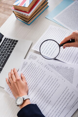 partial view of translator holding magnifier above documents near laptop and dictionaries of...