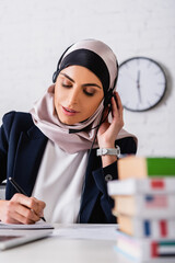 arabian translator in headset writing near dictionaries of foreign languages on blurred foreground