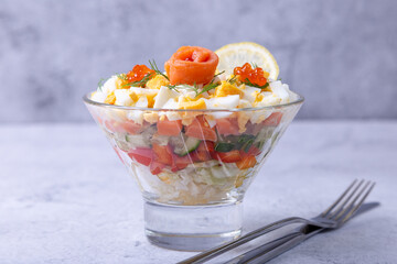 Layered salad with salted trout (salmon), caviar, eggs and vegetables. Traditional dish, one portion in a glass goblet on a gray background. Close-up.