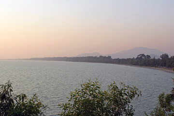 Landscape view over trees across ocean to the beach and shore with distant mountains and a clear sunset sky, southeast Asia