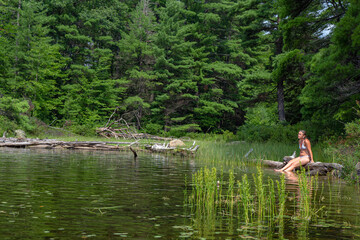 Fototapeta na wymiar Girl sitting on log in lake while camping in Canadian Provincial Park - Algonquin Number One Lake