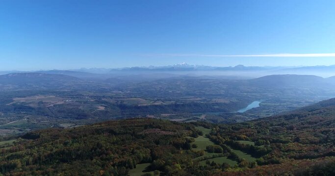 Aerial drone footage advancing toward a distant mountain range, with layered hills and a hazy valley below. A dramatic wide shot capturing the scale of the Alps and the surrounding landscape under cle