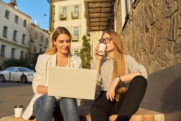 Two young smiling women looking at laptop monitor, sitting outdoors