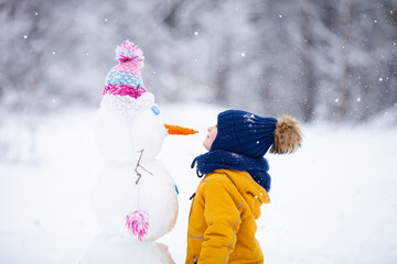 Little boy wearing bright knitted clothes looking at a snowman's nose on winter snowy day.  Love winter, bright colors and active outdoors leisure concept.