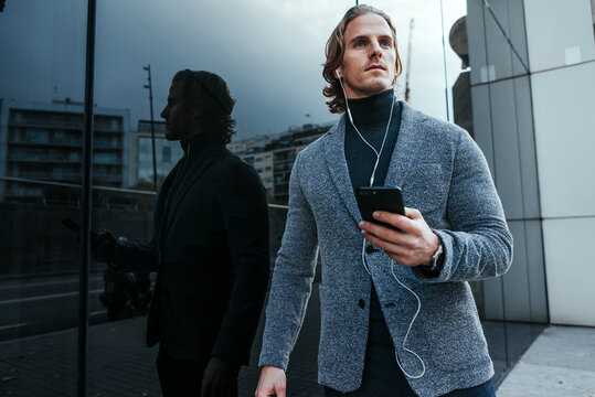 Modern businessman with smartphone on a city street. Handsome man wearing earphones and speaking on mobile phone, listening music or podcast outdoors.