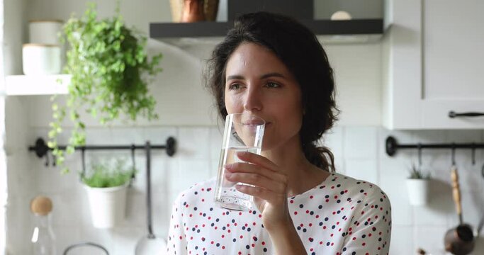 Head shot beautiful young dreamy caucasian woman drinking glass of fresh pure water, enjoying healthy morning habit alone in modern kitchen, starting new energetic day, feeling refreshed and hydrated.