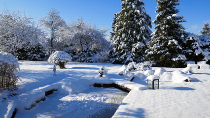 Gartenanlage im sonnigen Winter mit Schnee und gefrorenem Teich und schneebedeckten Bäumen