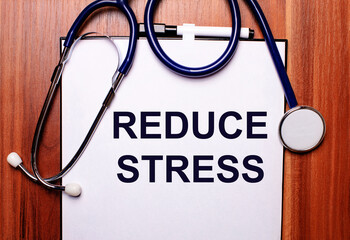 The word REDUCE STRESS is written on white paper on a wooden background near a stethoscope and black-framed glasses. Medical concept