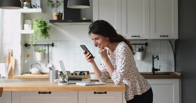 Addicted to social networks young happy beautiful woman scrolling profile pages, zooming photos, reading friends life news feed posts using mobile phone application, standing in modern kitchen alone.