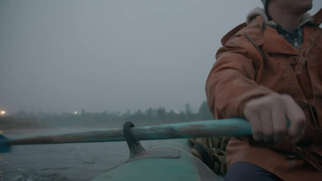 Close-up: the hand of a young man in an old orange raincoat rowing an inflatable boat in the early gray foggy morning. The guy in the raincoat works with his hands. Cold, dawn, loneliness. Slow mo, 4K