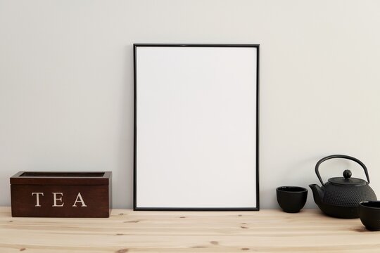 Kitchen wall art frame mockup, empty black frame on kitchen table, tea pot and cups.