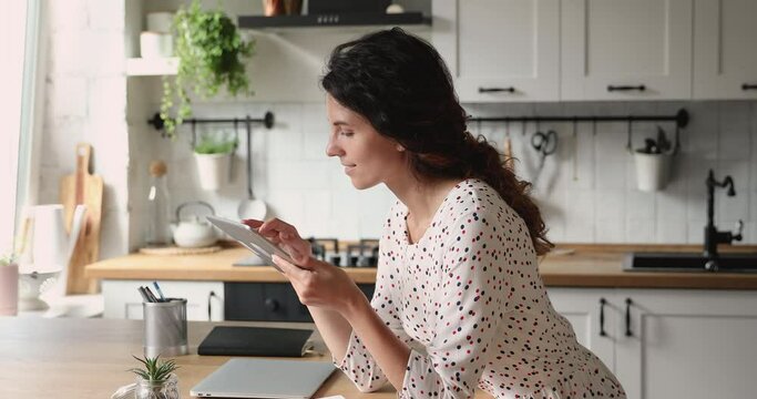 Smiling attractive young caucasian woman involved in digital computer tablet usage, web surfing information online, choosing goods in internet store, shopping or communicating with friends distantly.