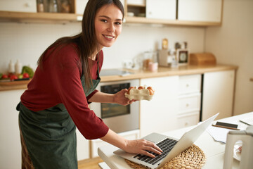 Attractive young female in apron holding carton box, going to make scrambled eggs, communicating online via messenger app using portable computer. Cute girl enjoying cooking in the morning