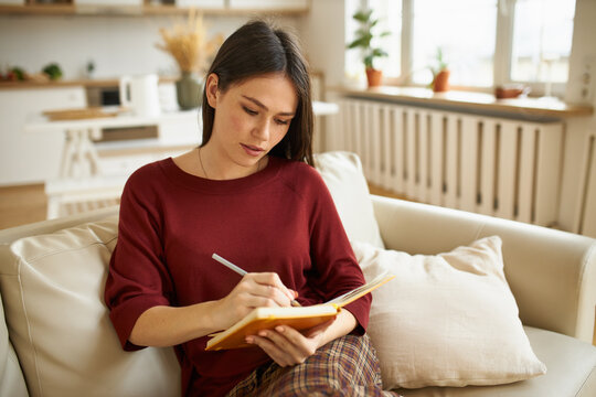 Picture of charming young female with dark haired sitting on comfortable couch holding pen, handwriting in copybook, making list of food products before grocery shopping. People and lifestyle