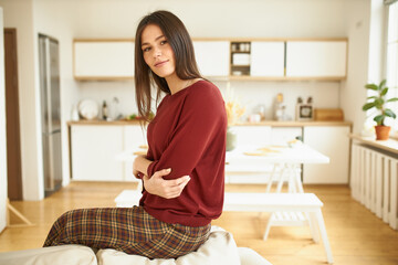 People, home, domesticity, comfort and relaxation concept. Adorable teenage girl in checkered trousers sitting on couch smiling at camera, spending lazy morning indoors, feeling comfortable