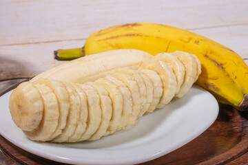 Sliced banana on a white plate on a wooden tray with another banana in the background