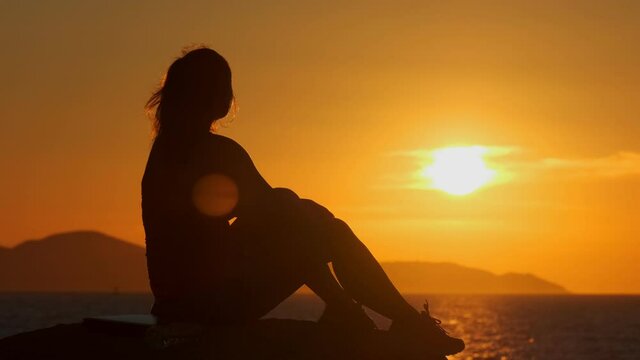 Silhouette of girl admiring beautiful sunset sitting on stone on sea coast.