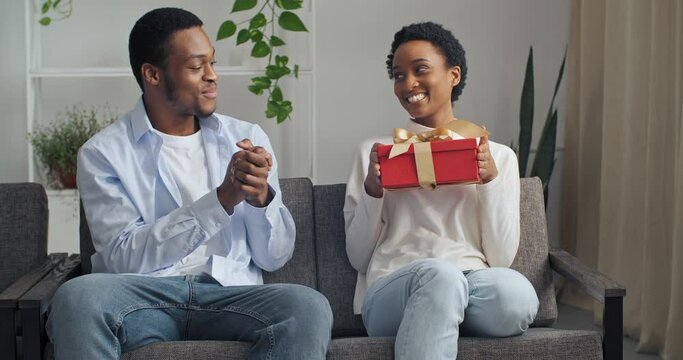 Beautiful young Afro American couple sitting on couch at home. Handsome black man guy giving his girlfriend atractive woman ethnic lady red gift box present congratulates happy anniversary birthday