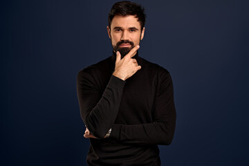 Stylish serious-looking young man with bristle in trendy high neck sweater, touching chin, rubbing beard and smirk as judging something, thinking or pondering checking out something interesting