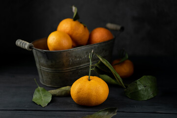 Several ripe tangerines with leaves in a metal container lie on a black table against a dark background. Rustic style with copyspace.