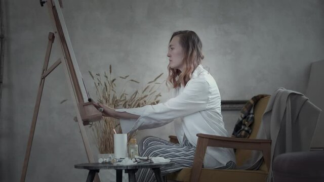 Side view of pacified young woman wearing white shirt sitting in yellow armchair draws at easel alone in a workshop or at home