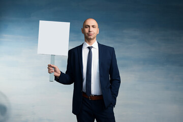 A bald man in a business blue suit spectacle and tie stands with holding a poster in his hand on a blue background
