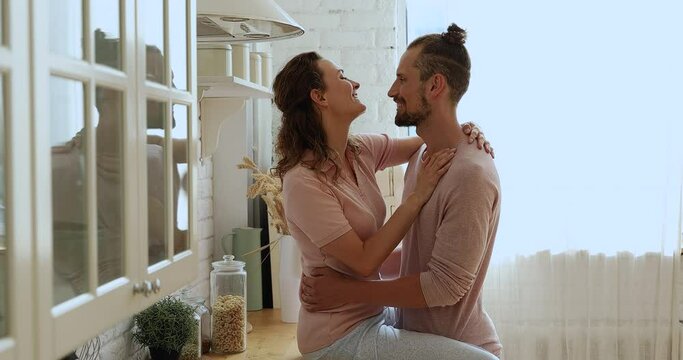 Beautiful couple enjoy romantic date in modern domestic kitchen. Happy wife sit on countertop cuddling beloved husband, affectionate spouses touch noses, kissing in lips feeling attraction and love