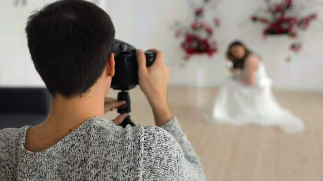 A male photographer with a camera takes pictures of a bride in a wedding dress indoors against a background of red live flowers
