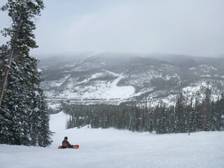 A snowboarder sitting on the slopes of a ski resort on a cloudy winter day, with snow covered trees around
