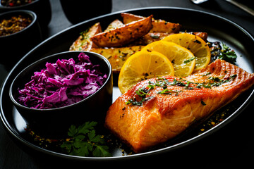 Fried salmon fillet with fried potatoes, red cabbage and lemon served on black plate on wooden table