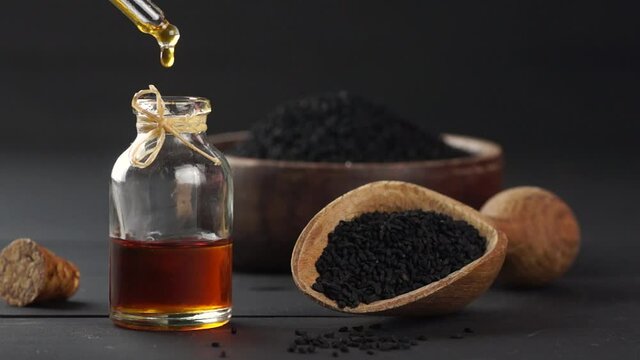 Glass bottle of black cumin seed essential oil , Nigella Sativa in scoop on black wooden background, oil dripping