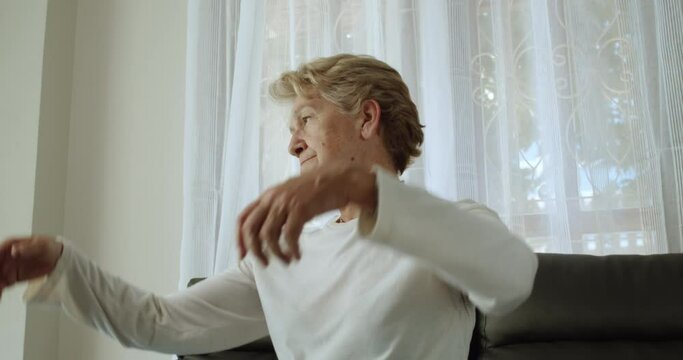 An elderly woman after retirement cut short hair with light brown hair wearing a long sleeve T-shirt and a necklace, feel bored and fatigue raised both arms to twist at her home alone.
