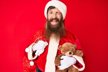 Handsome young red head man with long beard wearing santa claus costume holding teddy bear winking looking at the camera with sexy expression, cheerful and happy face.
