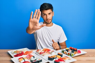 Young handsome man eating sushi sitting on the table doing stop sing with palm of the hand. warning...