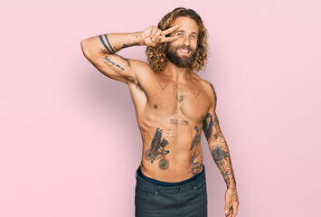Handsome man with beard and long hair standing shirtless showing tattoos doing peace symbol with...
