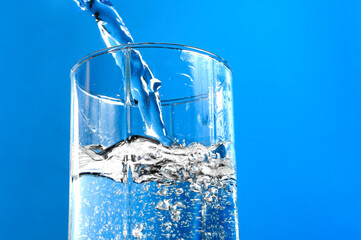 Pouring water into glass on blue background. Stream of clean water flowing in a glass.
