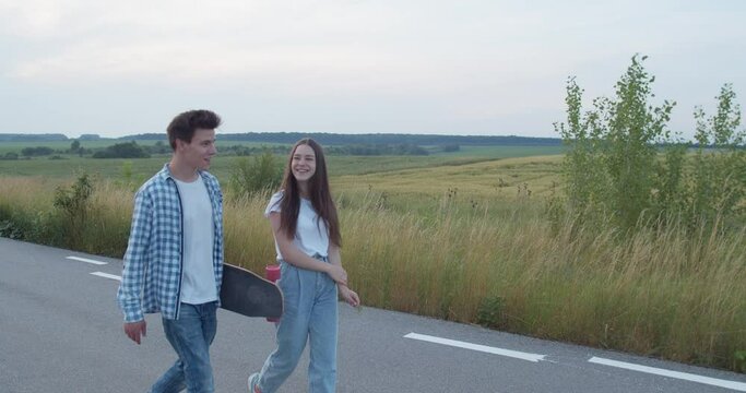 Two joyful friends walking with skateboard on road and having small talk