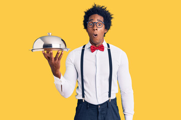 Handsome african american man with afro hair wearing waiter uniform holding silver tray scared and...
