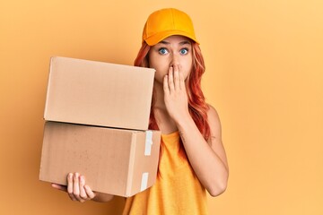 Young redhead woman holding delivery package covering mouth with hand, shocked and afraid for mistake. surprised expression
