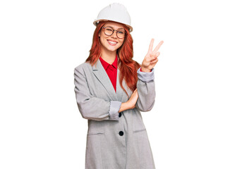 Young redhead woman wearing architect hardhat smiling with happy face winking at the camera doing victory sign. number two.