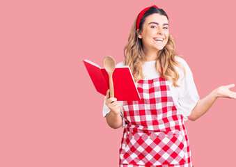 Young caucasian woman wearing apron holding recipe book and spoon celebrating victory with happy smile and winner expression with raised hands