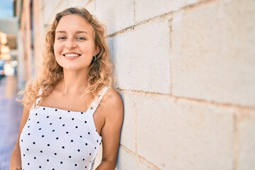 Young beautiful caucasian woman with blond hair smiling happy outdoors on a summer day leaning on the wall