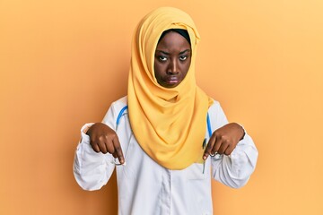 Beautiful african young woman wearing doctor uniform and hijab pointing down looking sad and upset, indicating direction with fingers, unhappy and depressed.