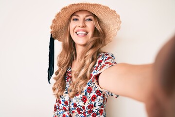 Young caucasian woman with blond hair wearing summer hat taking a selfie looking positive and happy standing and smiling with a confident smile showing teeth