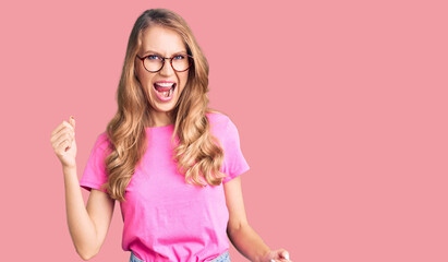 Young beautiful caucasian woman with blond hair wearing casual clothes and glasses angry and mad raising fist frustrated and furious while shouting with anger. rage and aggressive concept.