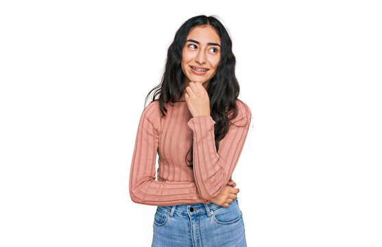 Hispanic teenager girl with dental braces wearing casual clothes with hand on chin thinking about question, pensive expression. smiling with thoughtful face. doubt concept.