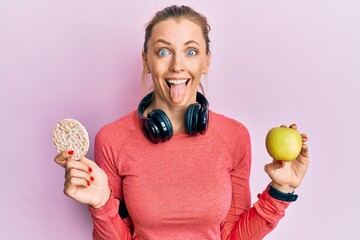 Beautiful caucasian sports woman holding green apple and rice crackers sticking tongue out happy with funny expression.